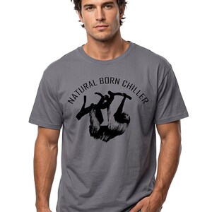 Pu&ograve; includere: T-shirt grigia con grafica nera di un bradipo appeso a un ramo. Il testo "NATURAL BORN CHILLER" &egrave; sopra il bradipo. La maglietta ha maniche corte e girocollo.