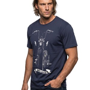 Puede incluir: Camiseta azul marino con un gr&aacute;fico detallado de una motocicleta, rayos y el texto "BAD SEED" en una pancarta. La camiseta se combina con vaqueros azules.