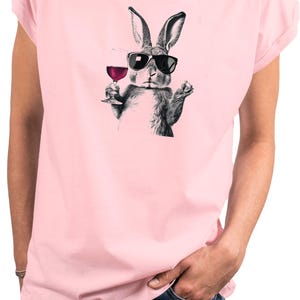 May include: Pink t-shirt featuring a black and white illustration of a rabbit wearing sunglasses and holding a glass of red wine. The rabbit is the central focus, with the wine glass and sunglasses adding detail to the design. The shirt is a solid pink color.