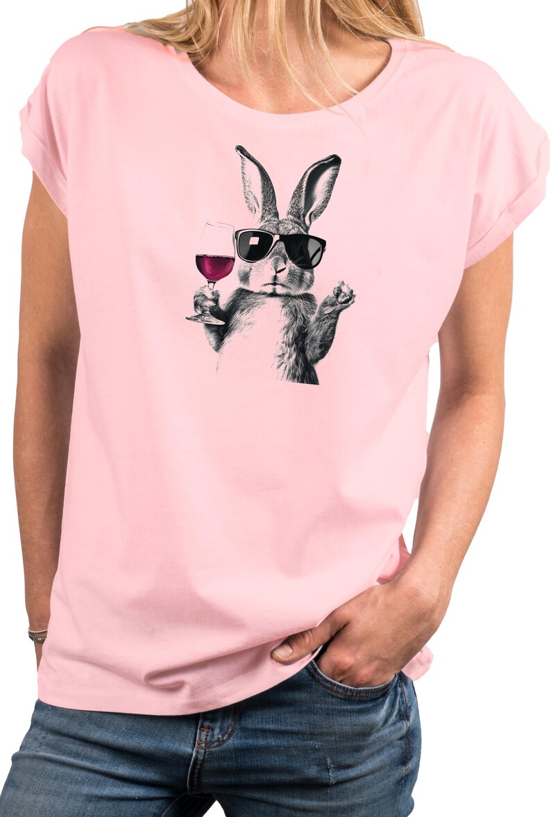 May include: Pink t-shirt featuring a black and white illustration of a rabbit wearing sunglasses and holding a glass of red wine. The rabbit is the central focus, with the wine glass and sunglasses adding detail to the design. The shirt is a solid pink color.