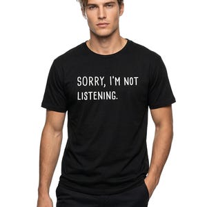 May include: Black crew neck t-shirt with the text "SORRY, I'M NOT LISTENING" in white. The person is wearing black pants and has their hand in their pocket.