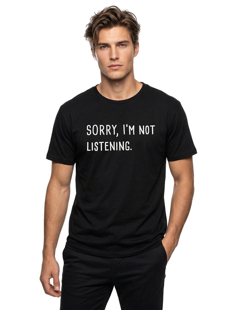 May include: Black crew neck t-shirt with the text "SORRY, I'M NOT LISTENING" in white. The person is wearing black pants and has their hand in their pocket.