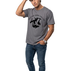 Pu&ograve; includere: T-shirt grigio con la scritta "NATURAL BORN CHILLER" e una grafica di bradipo nero. La persona indossa jeans blu e scarpe da ginnastica bianche. Un orologio d'argento &egrave; al polso.