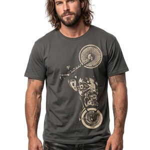 以下が含まれることがあります： 濃いグレーのTシャツ。ベージュのバイクのグラフィックが特徴です。デザインには、バイクの前輪と後輪、エンジン部品、フレームが含まれています。Tシャツは、クラシックなクルーネックです。
