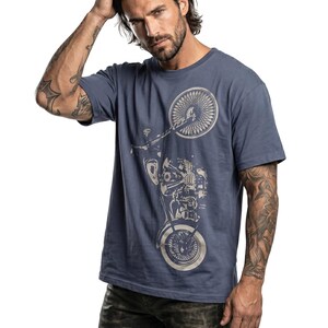 以下が含まれることがあります： 明るい色合いのバイクのグラフィックが特徴の青いTシャツ。Tシャツは半袖とクルーネックです。モデルは濃い色のジーンズを着用しています。背景は白です。