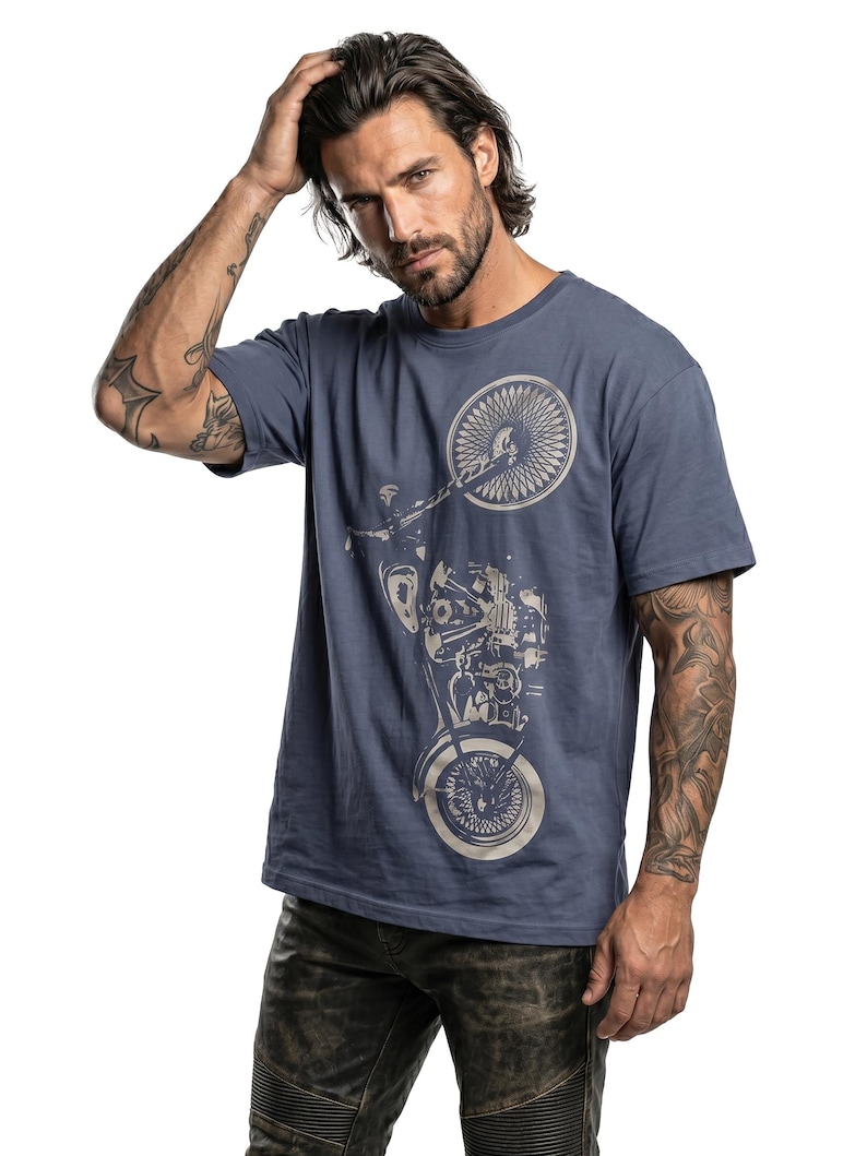 以下が含まれることがあります： 明るい色合いのバイクのグラフィックが特徴の青いTシャツ。Tシャツは半袖とクルーネックです。モデルは濃い色のジーンズを着用しています。背景は白です。