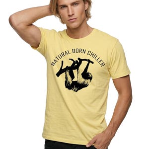 Pu&ograve; includere: T-shirt giallo chiaro con la scritta "NATURAL BORN CHILLER" in un design circolare. La maglietta presenta una grafica nera di un bradipo. Il modello indossa pantaloni neri.