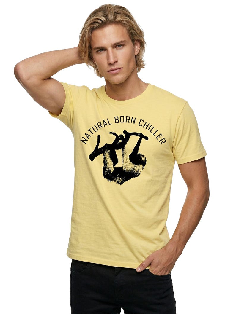 Pu&ograve; includere: T-shirt giallo chiaro con la scritta "NATURAL BORN CHILLER" in un design circolare. La maglietta presenta una grafica nera di un bradipo. Il modello indossa pantaloni neri.