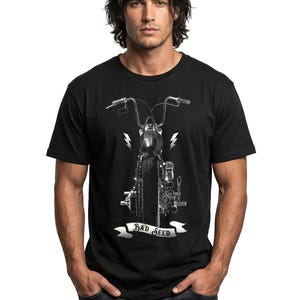 Puede incluir: Camiseta negra con un gr&aacute;fico de motocicleta en escala de grises, que muestra el manillar y rayos. El texto "BAD SEED" est&aacute; en una pancarta. El modelo lleva vaqueros azules. La imagen est&aacute; sobre un fondo blanco.