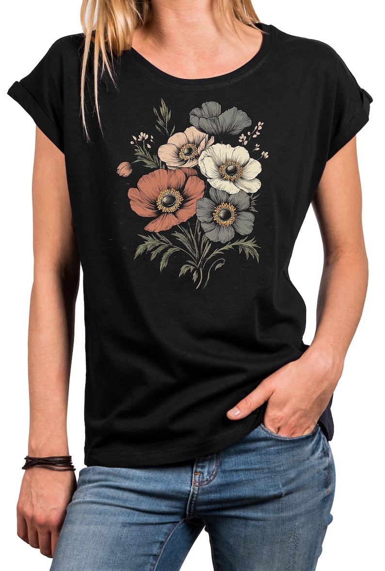May include: Black t-shirt featuring a floral design with peach, white, and gray anemone flowers. The flowers have yellow centers and are arranged with green leaves and stems. The shirt has rolled-up sleeves and a round neckline.