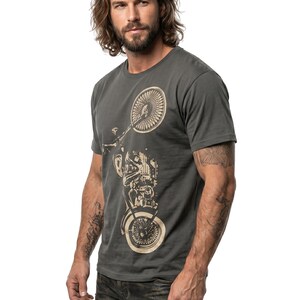 以下が含まれることがあります： ベージュのオートバイのグラフィックが特徴の濃い灰色のTシャツ。半袖シャツは、長い茶色の髪と腕にタトゥーのある人が着用しています。その人は濃い灰色のジーンズを履いています。
