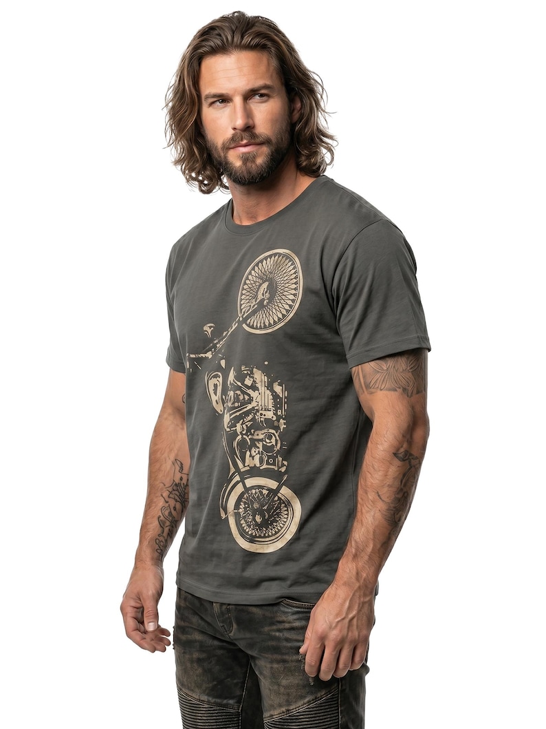 以下が含まれることがあります： ベージュのオートバイのグラフィックが特徴の濃い灰色のTシャツ。半袖シャツは、長い茶色の髪と腕にタトゥーのある人が着用しています。その人は濃い灰色のジーンズを履いています。