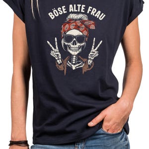 K&ouml;nnte beinhalten: Marineblaues T-Shirt mit einem Skelett-Motiv, das eine Sonnenbrille und ein rotes Bandana tr&auml;gt und Friedenszeichen zeigt. Der Text "B&Ouml;SE ALTE FRAU" ist &uuml;ber dem Motiv aufgedruckt. Das T-Shirt hat hochgekrempelte &Auml;rmel.