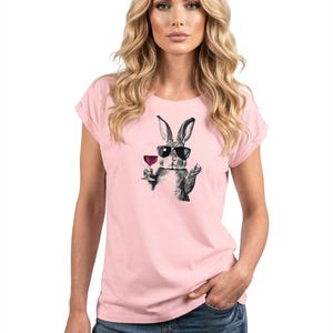 May include: A light pink t-shirt featuring a black and white illustration of a rabbit wearing sunglasses and holding a glass of wine. The rabbit graphic is centered on the shirt. The shirt has rolled-up sleeves.
