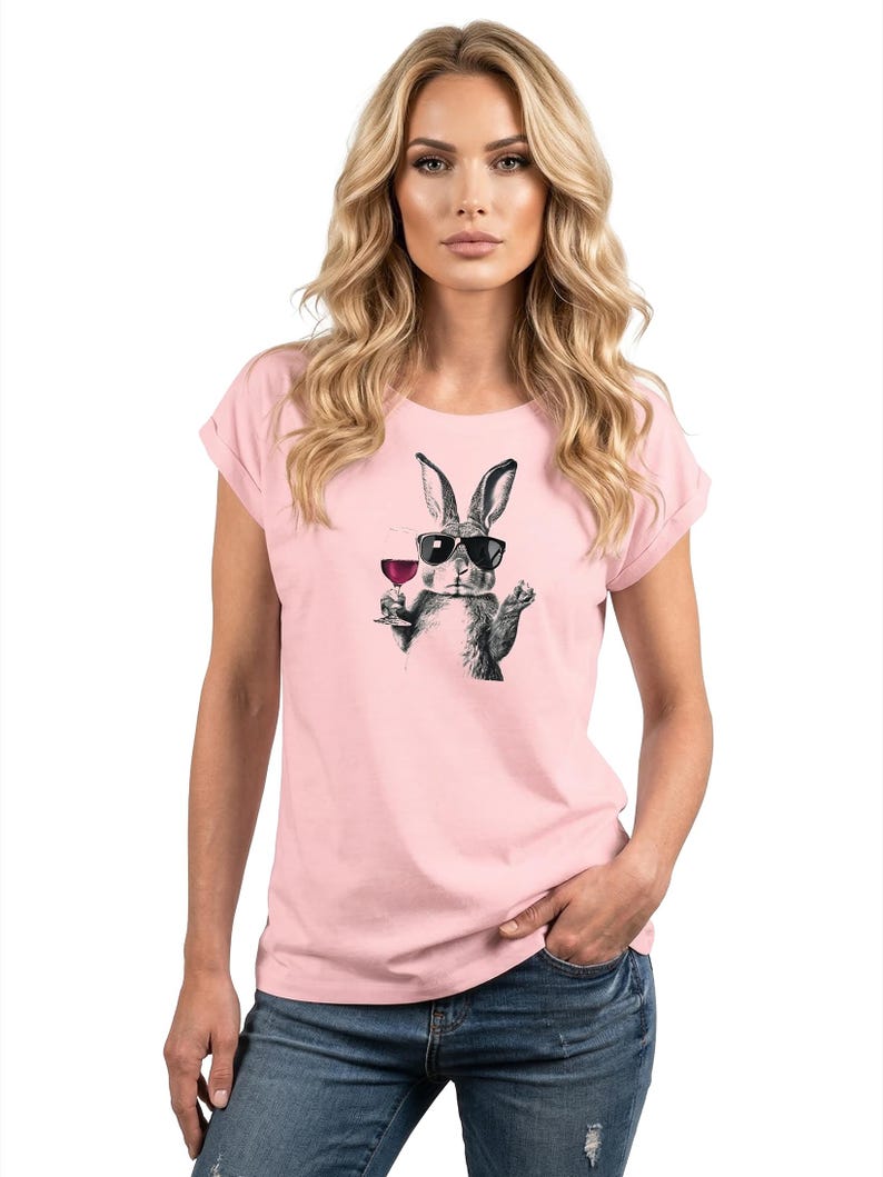 May include: A light pink t-shirt featuring a black and white illustration of a rabbit wearing sunglasses and holding a glass of wine. The rabbit graphic is centered on the shirt. The shirt has rolled-up sleeves.