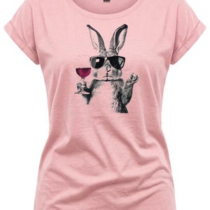 May include: A light pink t-shirt featuring a black and white illustration of a rabbit wearing sunglasses and holding a glass of red wine. The shirt has rolled-up sleeves and a crew neck.