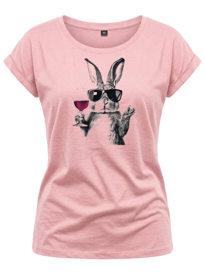 May include: A light pink t-shirt featuring a black and white illustration of a rabbit wearing sunglasses and holding a glass of red wine. The shirt has rolled-up sleeves and a crew neck.
