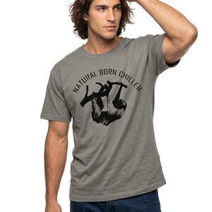 Pu&ograve; includere: T-shirt grigio con la scritta "NATURAL BORN CHILLER" e una grafica di bradipo nero. La maglietta &egrave; a girocollo e maniche corte. Il modello indossa jeans blu.