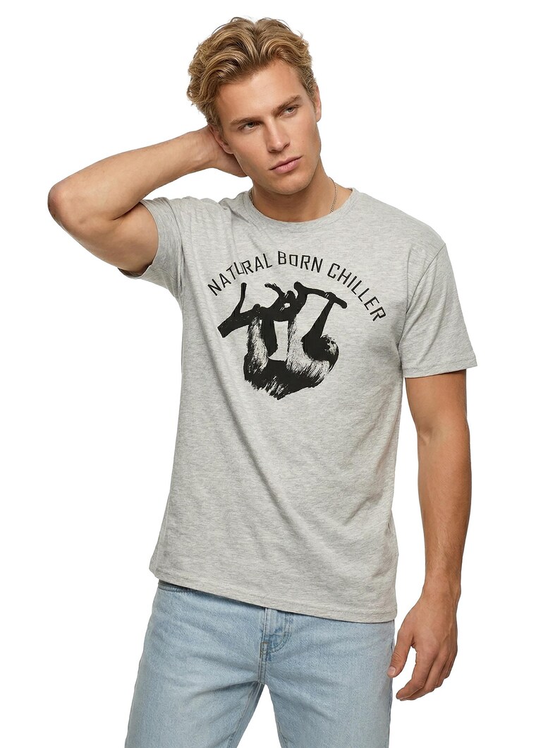 Pu&ograve; includere: T-shirt grigio chiaro con la scritta "NATURAL BORN CHILLER" e una grafica di bradipo nero. La maglietta &egrave; a maniche corte e ha un girocollo. Il modello indossa jeans azzurri.
