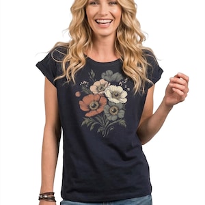 May include: Navy blue t-shirt featuring a floral design in shades of peach, white, and gray. The t-shirt has rolled sleeves and is paired with blue jeans. A brown leather bracelet is visible on the left wrist.