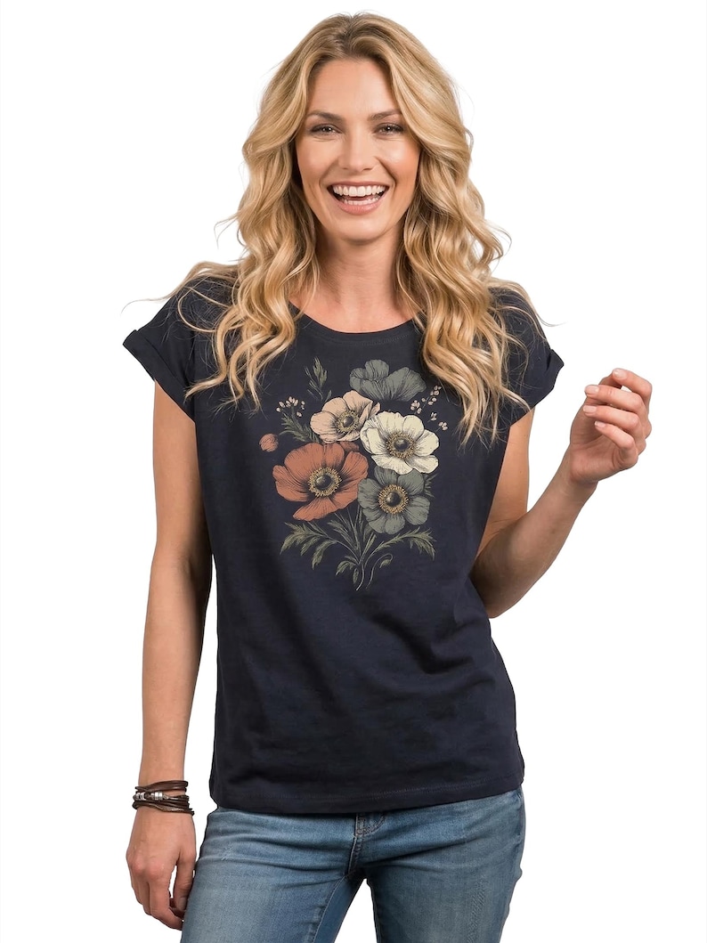 May include: Navy blue t-shirt featuring a floral design in shades of peach, white, and gray. The t-shirt has rolled sleeves and is paired with blue jeans. A brown leather bracelet is visible on the left wrist.