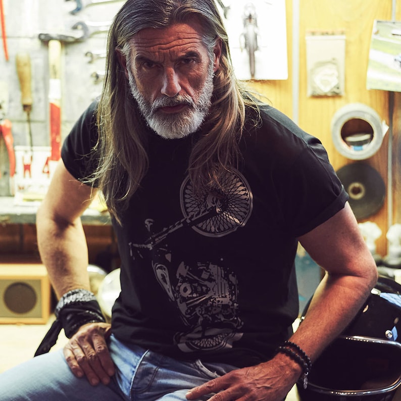 以下が含まれることがあります： バイクのグラフィックがプリントされた黒いTシャツ。青いジーンズと黒いリストバンドを着用。長い灰色の髪とひげを生やしている。背景には工具や設備が写っている。
