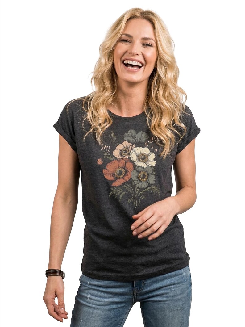 May include: Dark gray t-shirt featuring a floral design with orange, white, and gray flowers. The short-sleeved tee is paired with blue jeans. A brown leather bracelet is worn on the left wrist.