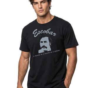 May include: Black t-shirt featuring a gray portrait and the name "Escobar" above the text "A FRIEND OF A FRIEND OF A FRIEND". The shirt is a crew neck and is being worn.