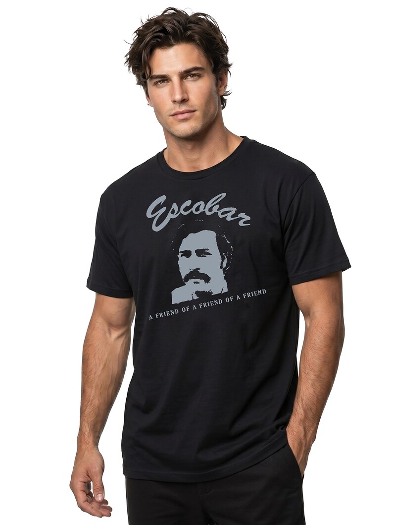 May include: Black t-shirt featuring a gray portrait and the name "Escobar" above the text "A FRIEND OF A FRIEND OF A FRIEND". The shirt is a crew neck and is being worn.