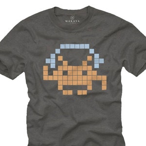 Op de afbeelding: Donkergrijs t-shirt met een gepixeld alien-ontwerp in oranje en lichtblauw. Het ontwerp doet denken aan klassieke arcadespellen. Het shirt heeft een ronde hals en korte mouwen. De merknaam "MAKAYA" staat bovenaan.