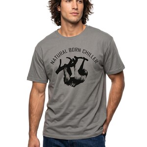 Pu&ograve; includere: T-shirt grigio con la scritta "NATURAL BORN CHILLER" e una grafica nera di un bradipo appeso a un ramo. La maglietta a maniche corte &egrave; abbinata a jeans blu.