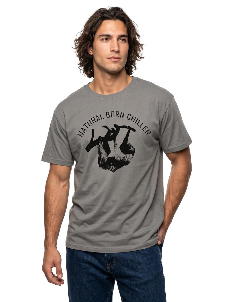 Pu&ograve; includere: T-shirt grigio con la scritta "NATURAL BORN CHILLER" e una grafica nera di un bradipo appeso a un ramo. La maglietta a maniche corte &egrave; abbinata a jeans blu.