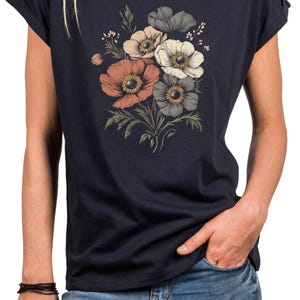 May include: Navy blue t-shirt featuring a floral design with orange, white, and gray anemone flowers. The flowers have yellow centers and green leaves. The shirt has rolled sleeves and a round neckline. The person is wearing blue jeans.