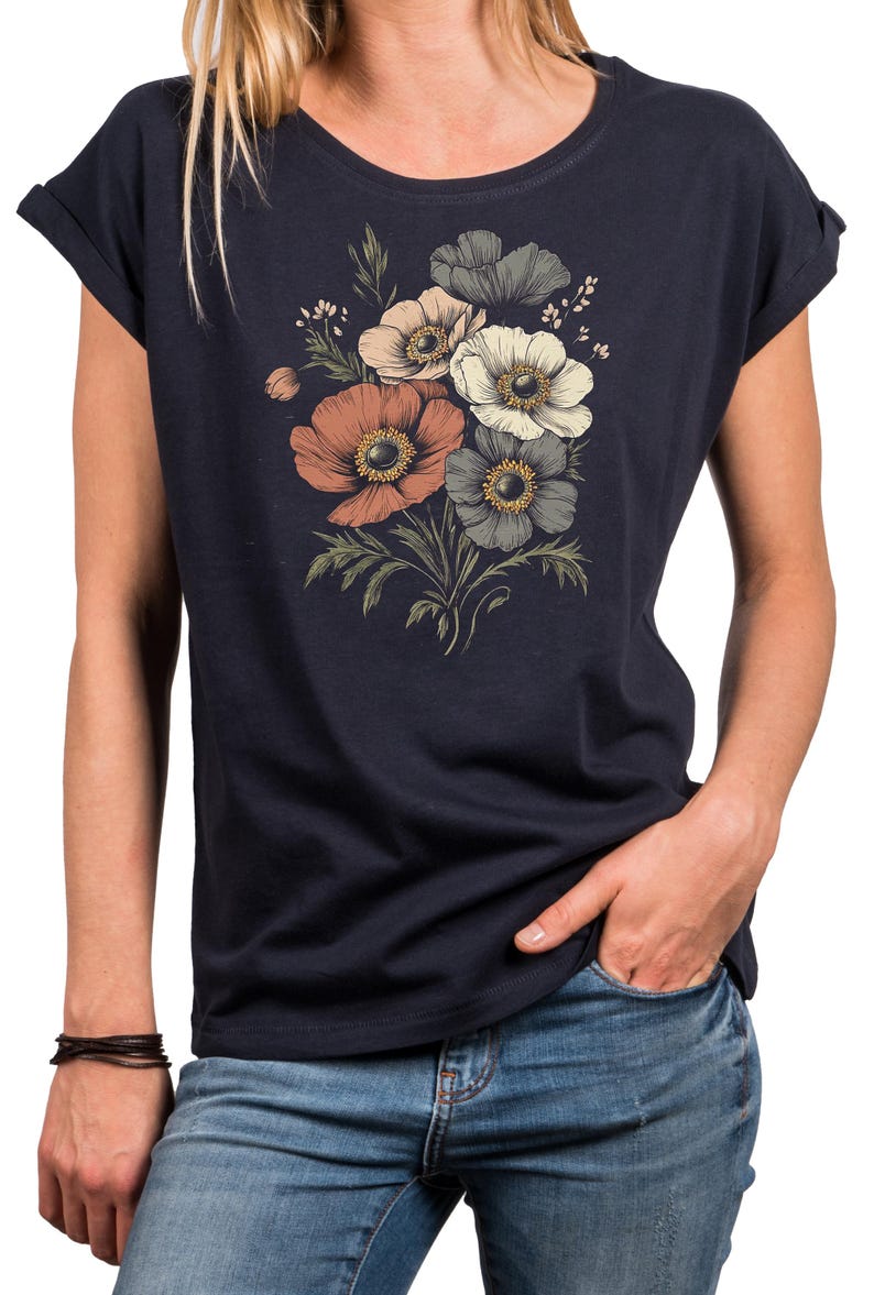 May include: Navy blue t-shirt featuring a floral design with orange, white, and gray anemone flowers. The flowers have yellow centers and green leaves. The shirt has rolled sleeves and a round neckline. The person is wearing blue jeans.