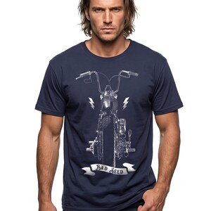 Puede incluir: Camiseta azul marino con una ilustraci&oacute;n detallada en escala de grises de la parte delantera de una motocicleta. El dise&ntilde;o incluye rayos y una pancarta que dice "BAD SEED". La camiseta la lleva una persona con pelo largo y casta&ntilde;o claro.