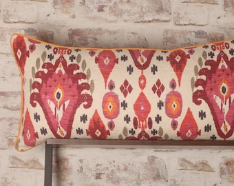 May include: A long, rectangular lumbar pillow with a cream-colored background and a colorful ikat pattern in shades of red, pink, orange, and green. The pillow has a yellow trim.