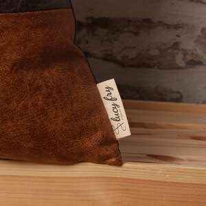 May include: A brown and black throw pillow with a brown suede-like texture. The pillow has a small white tag sewn on the side with the text "lucy fry" in cursive.