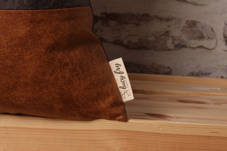 May include: A brown and black throw pillow with a brown suede-like texture. The pillow has a small white tag sewn on the side with the text "lucy fry" in cursive.