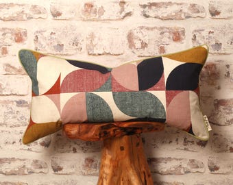 May include: A lumbar pillow with a geometric pattern of circles and semi-circles in shades of pink, blue, brown, and white. The pillow is sitting on a wooden stool.