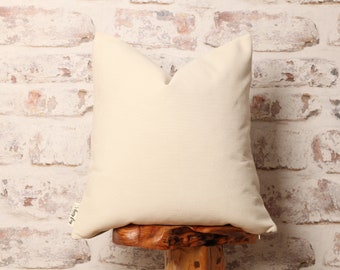 May include: A cream-colored pillow with a subtle ribbed texture. The pillow is sitting on a wooden stool.