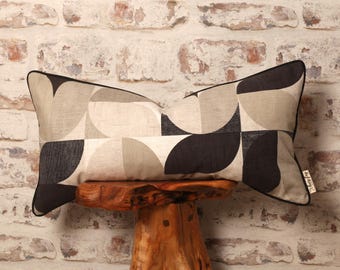 May include: Rectangular decorative pillow with a geometric pattern in shades of black, beige, and gray. The pillow has a black border and is displayed on a wooden stool. The pillow's design features curved shapes and a textured appearance.