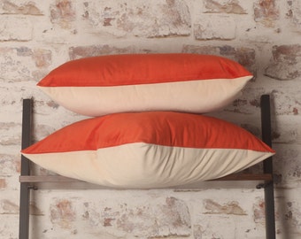 May include: Two decorative throw pillows with a cream colored base and a bright orange top. The pillows are stacked on a wooden shelf with black metal supports.