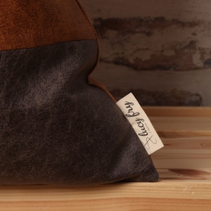 May include: A brown and gray throw pillow with a brown leather-like texture on the top half and a gray suede-like texture on the bottom half. The pillow has a white tag attached to it with the text "lucky frog" written in black.