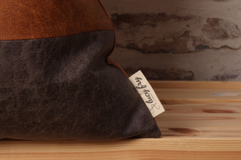 May include: A brown and gray throw pillow with a brown leather-like texture on the top half and a gray suede-like texture on the bottom half. The pillow has a white tag attached to it with the text "lucky frog" written in black.