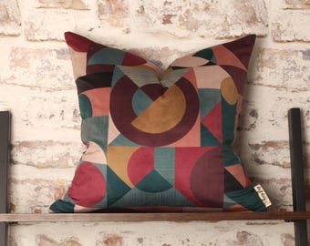 May include: A geometric patterned throw pillow with a burgundy, teal, and gold color scheme. The pillow is on a wooden shelf against a white brick wall.