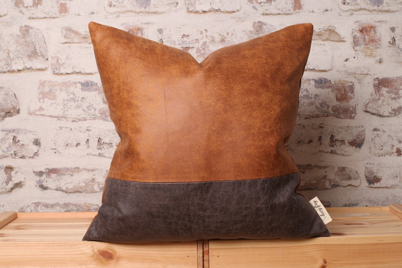 May include: A brown and gray faux leather pillow with a brown top and a gray bottom. The pillow has a small tag on the bottom right corner that says "Cozy Up".
