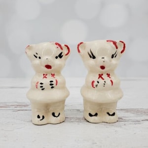 May include: A pair of vintage ceramic salt and pepper shakers shaped like bears. The bears are white with red and black details. The bears have a grumpy expression.