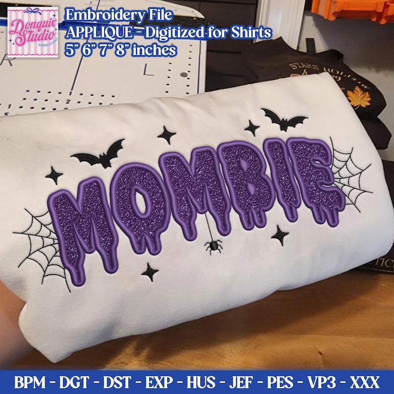 May include: A white fabric featuring the purple glittery embroidered text MOMBIE in a dripping font style. The design includes black embroidered spiderwebs, small bats, stars, and a spider hanging from the letter I. The text is digitized for shirts.