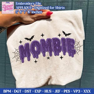 May include: A cream-colored t-shirt featuring a purple glittery applique design that reads MOMBIE in a dripping font. The design includes black embroidered spiderwebs, bats, stars, and a small spider. The file is digitized for 5, 6, 7, and 8 inch sizes.