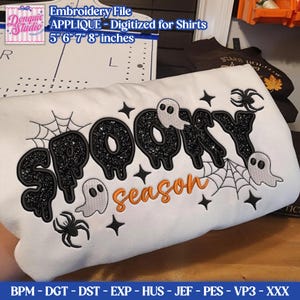 Peut inclure: Un tissu blanc avec un motif brodé comprenant le texte noir pailleté SPOOKY et le texte orange season. Le design inclut des fantômes blancs, des toiles d'araignée noires, des araignées et des étoiles. Le texte en haut indique Embroidery File APPLIQUE - Digitized for Shirts 5 6 7 8 inches.